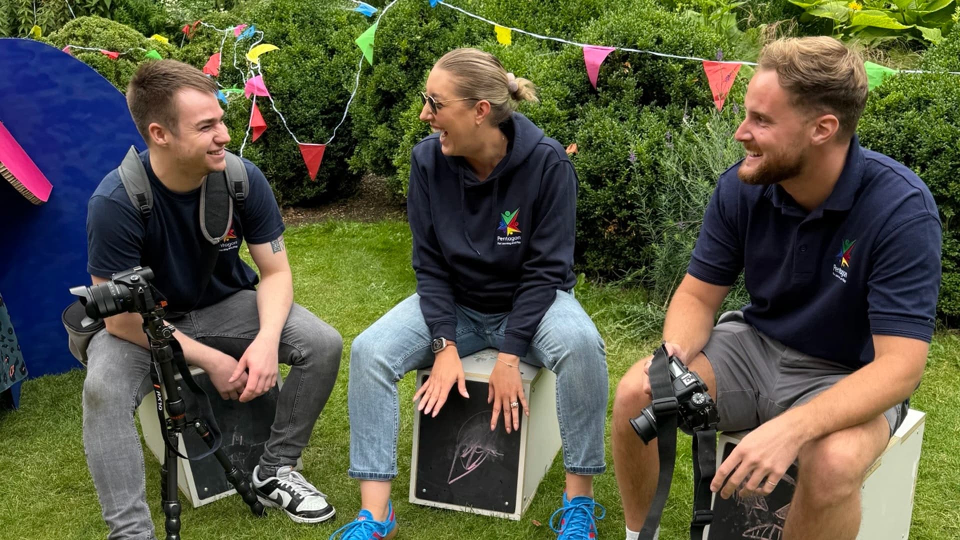 Three team members from Pentagon Play lauging and smiling whilst sat on musicial play equipment