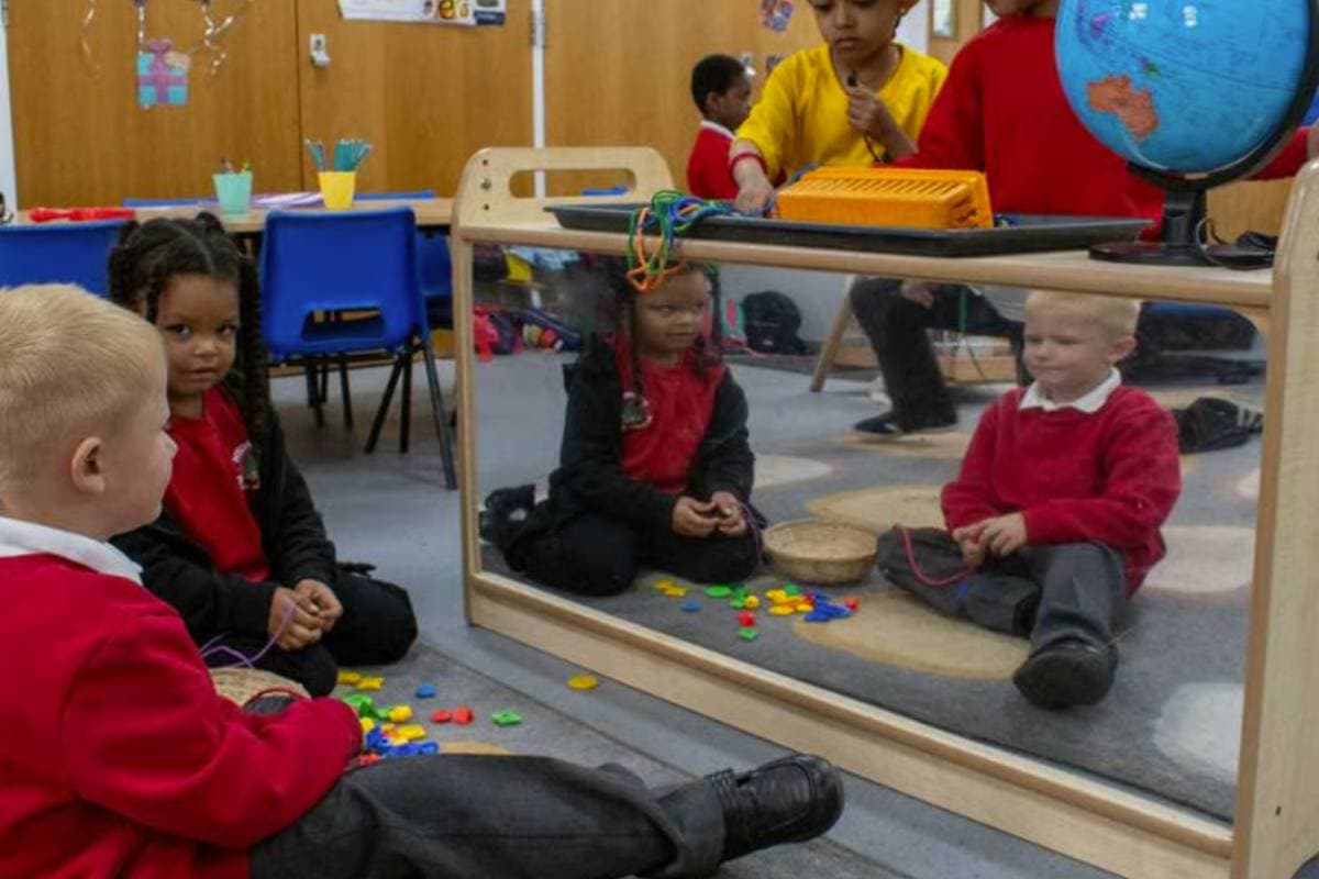 Two nursery children sitting on the floor in a classroom, looking in a mirror on the back of a bookcase.