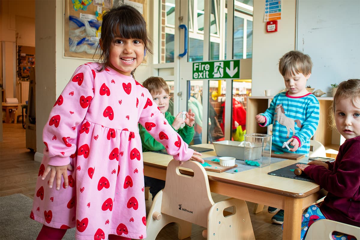 A toddler wearing a pink dress, standing against a wooden chair in a nursery