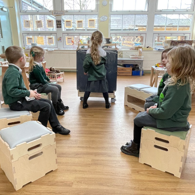 school children sat on stack and sit stools around art easel