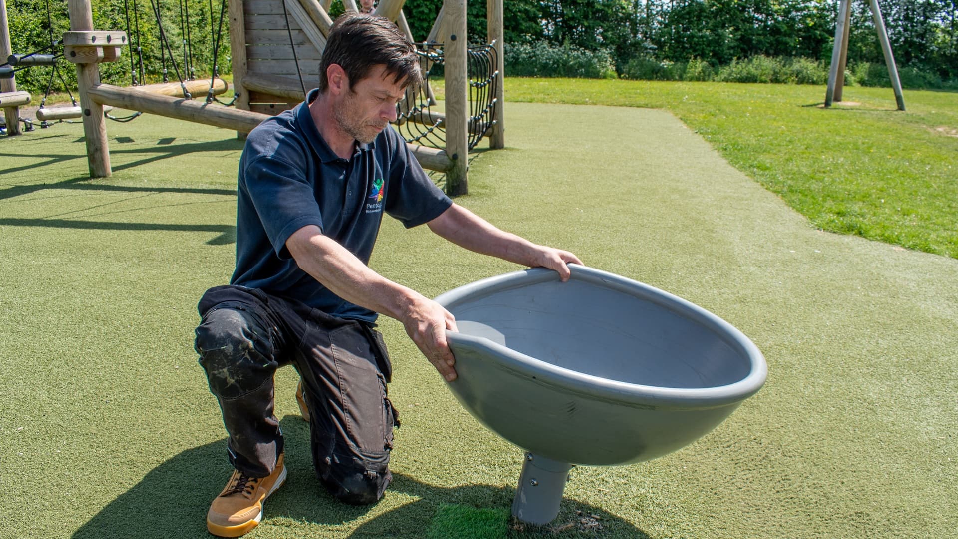 A playground inspector is holding a screwdriver as she prys away the wetpour that surrounds a wooden beam, showcasing the mold and rot that is affecting the piece of play equipment
